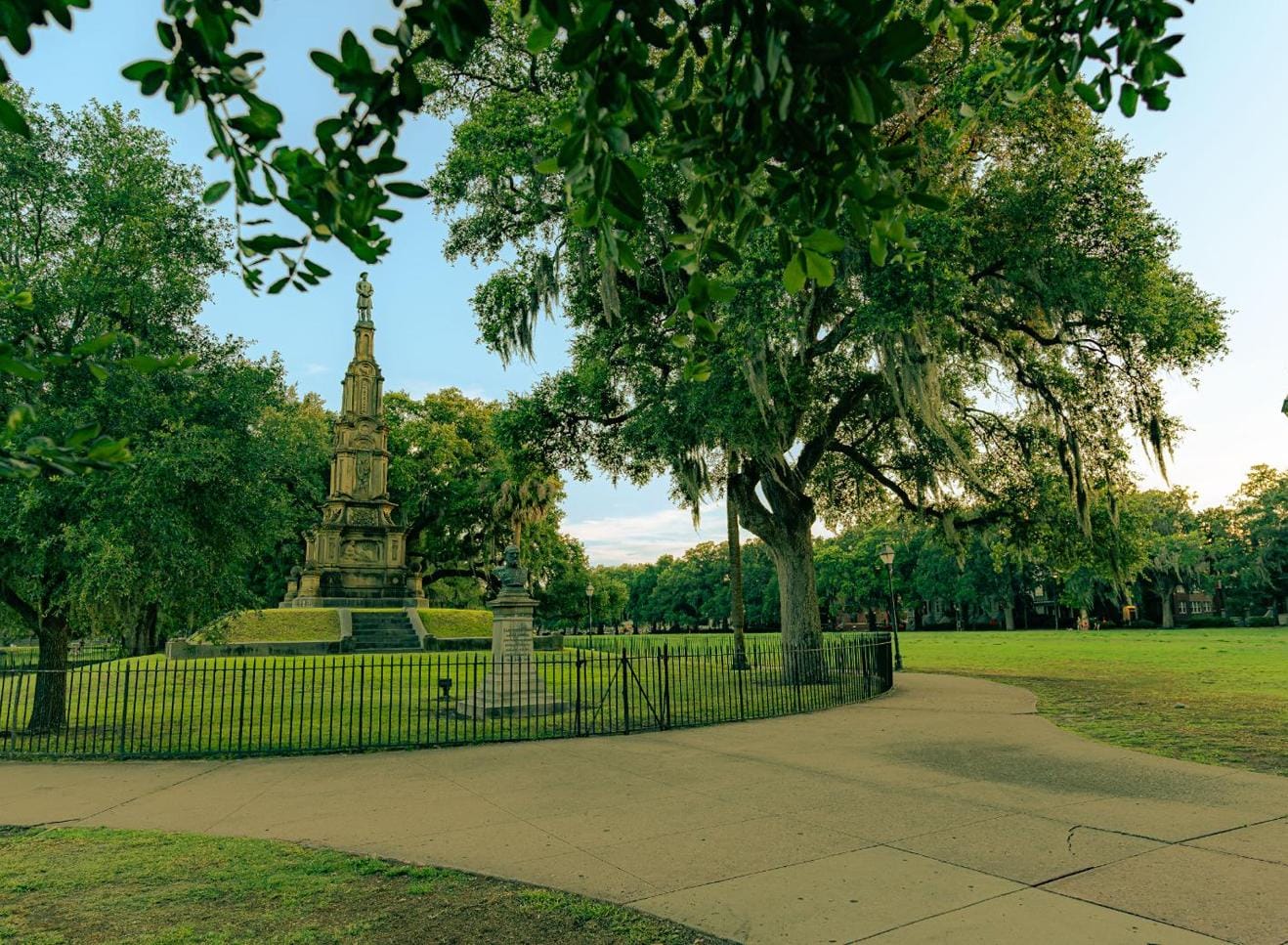 A tall monument stands in a grassy park surrounded by a black metal fence and large oak trees with Spanish moss.